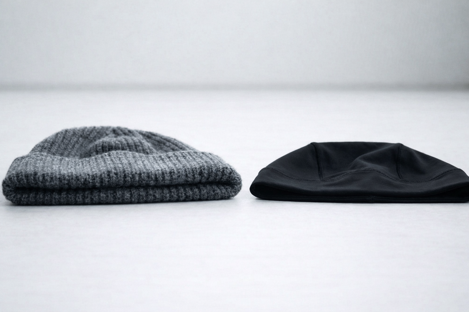 Two folded beanies placed side by side, a gray knitted beanie and a black smooth-fabric beanie, displayed on a white surface to highlight material and shape differences.