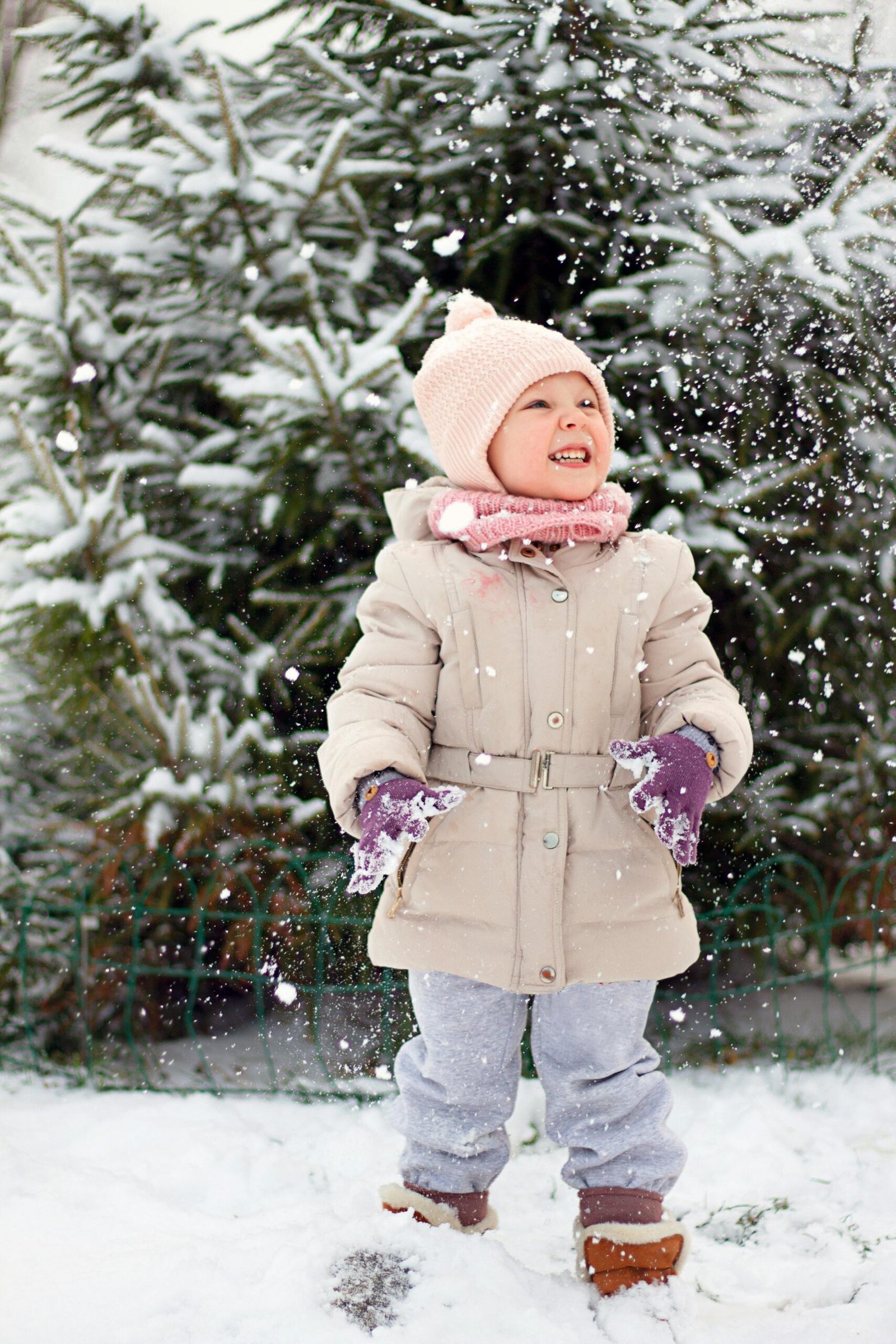 A young child wearing a warm winter coat, knitted beanie, scarf, and gloves playing in falling snow outdoors near snow-covered evergreen trees.