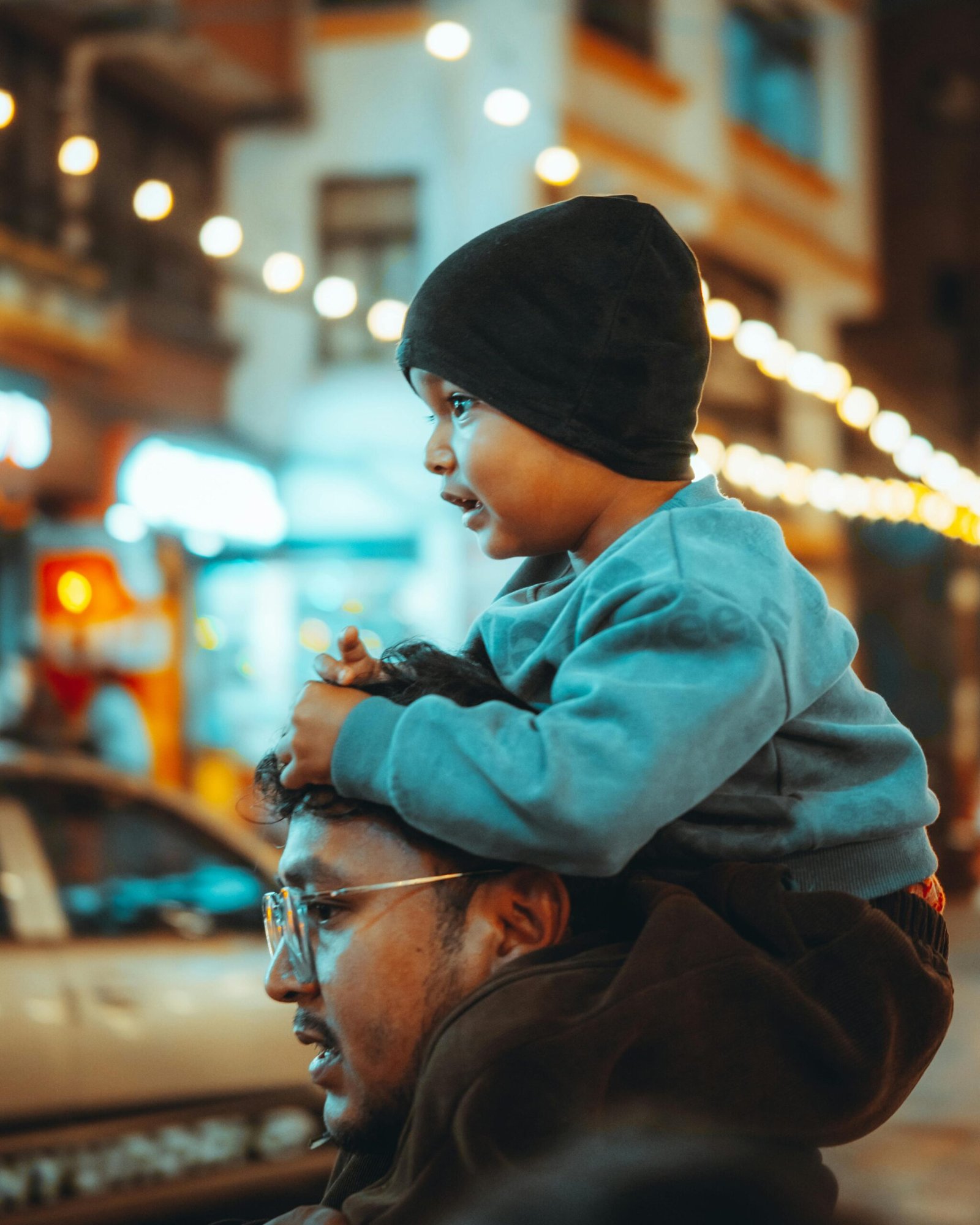 A child wearing a beanie sits on an adult’s shoulders in a brightly lit city street at night, with warm bokeh lights in the background